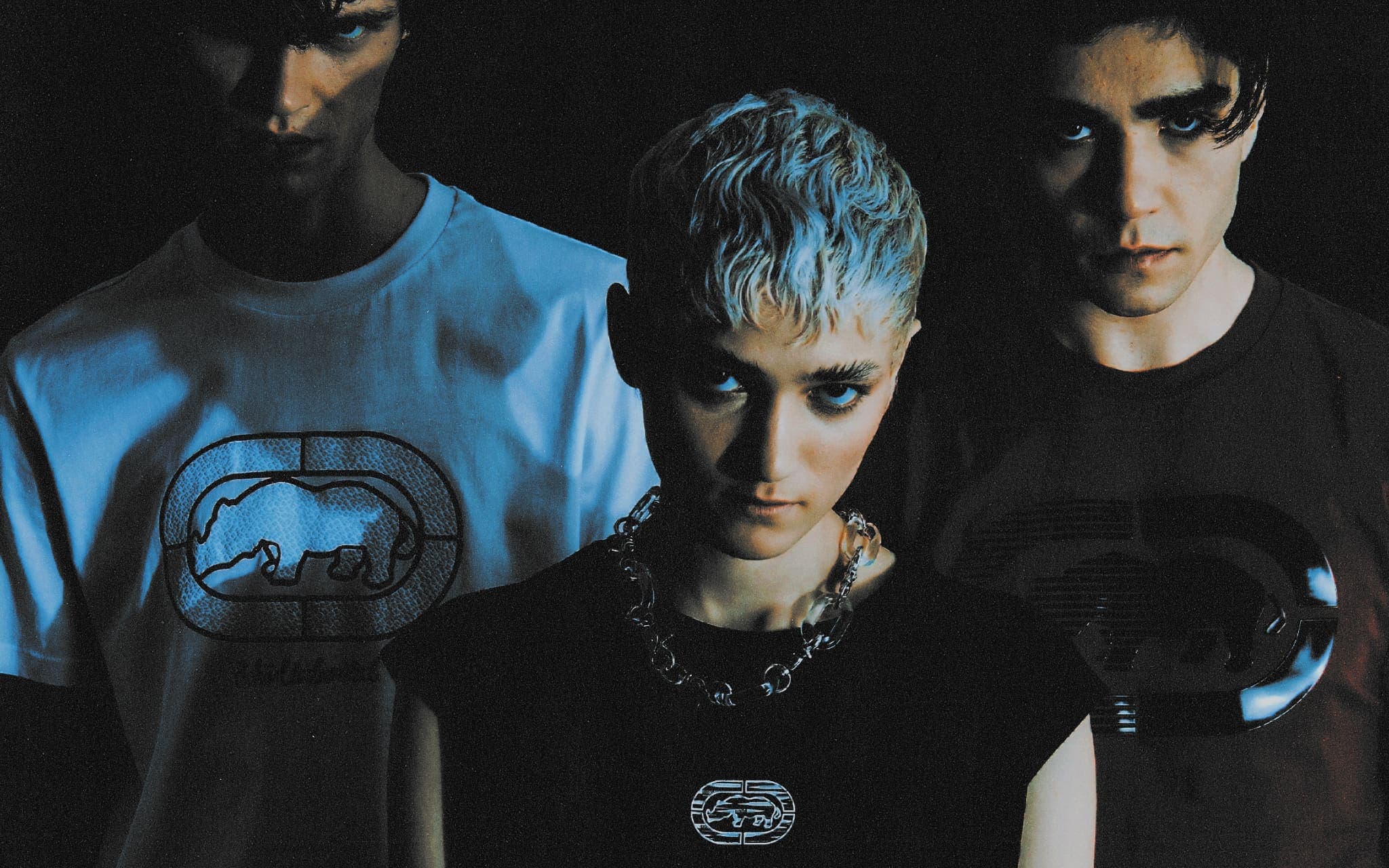 Three models in a dark studio wearing Ecko UNLTD tees, the rhino emblem on each chest: outlined on white, small and silver on black, large and glossy on brown