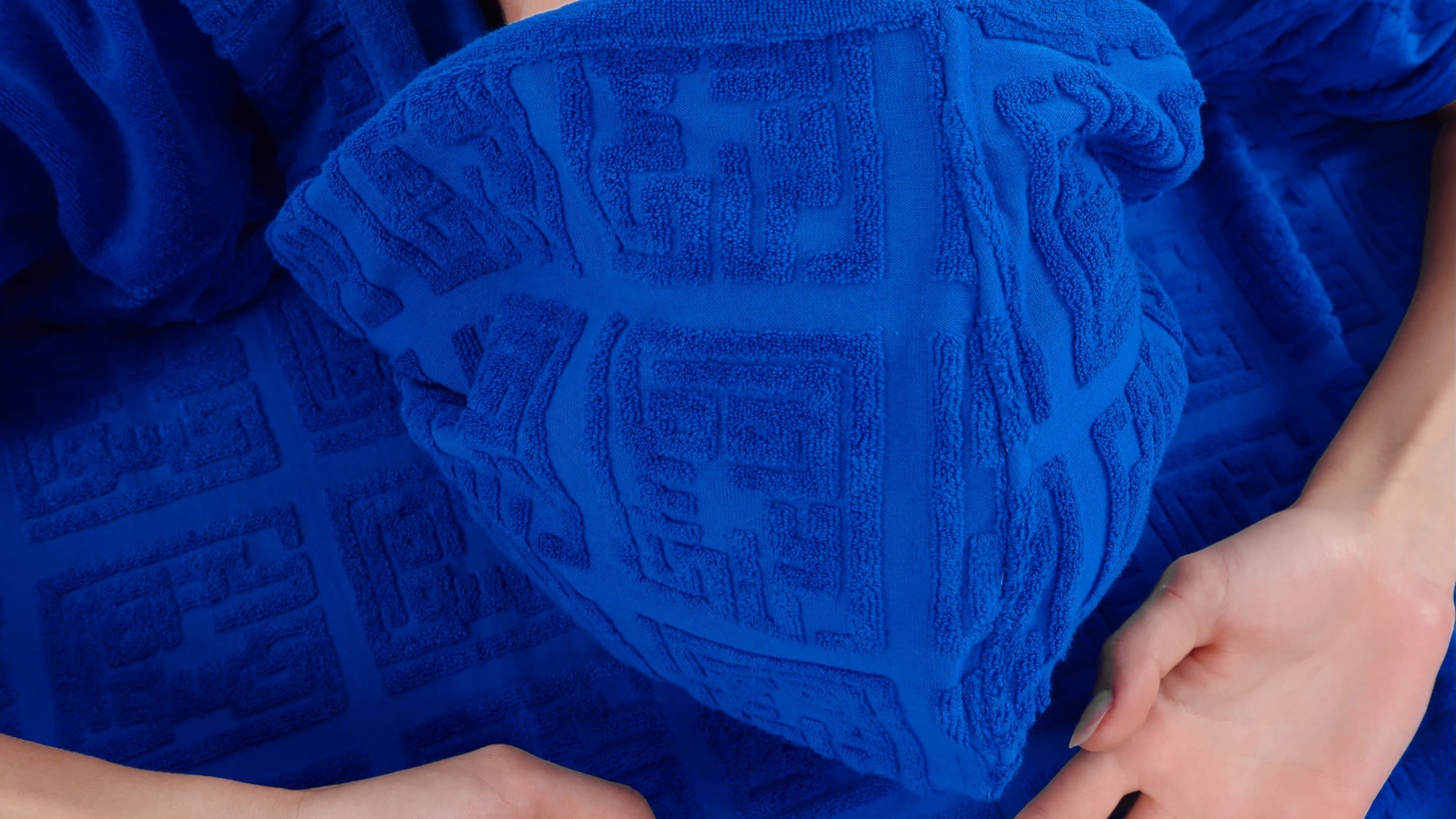 A blue terry hood with the kufic maze raised in the pile, pulled over the face, one hand open on the fabric
