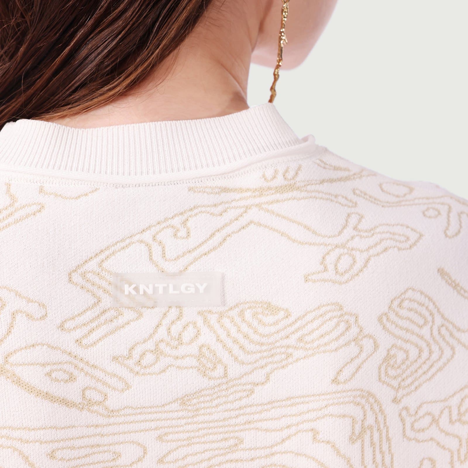 The back of a cream knit top with the organic line print knitted in gold thread and a KNTLGY label at the neck